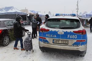 Policjanci na targowisku wręczają ulotki i elementy odblaskowe seniorom, z boku stoi radiowóz.