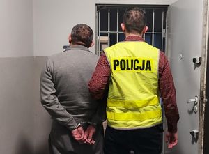 Policjant prowadzi zatrzymanego  po korytarzu do pomieszczenia dla osób zatrzymanych.