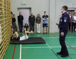 Sala gimnastyczna, na której rozłożony jest tor , jeden z zawodników wykonuje skłony z piłką, wokół stoją uczestnicu zawodów oraz policjant.