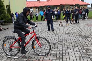 Na placu przed szkołą ustawiony tor przeszkód, uczestnik turnieju pokonuje przeszkody na rowerze, policjant ocenia wykonanie zadań.