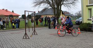 Na placu przed szkołą ustawiony tor przeszkód, uczestnik turnieju pokonuje przeszkody na rowerze, policjant ocenia wykonanie zadań.