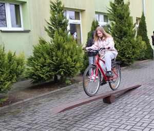 Na placu przed szkołą ustawiony tor przeszkód, uczestnik turnieju pokonuje przeszkody na rowerze.