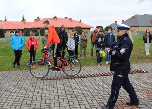 Na placu przed szkołą ustawiony tor przeszkód, uczestnik turnieju pokonuje przeszkody na rowerze, policjantka ocenia wykonanie zadań.