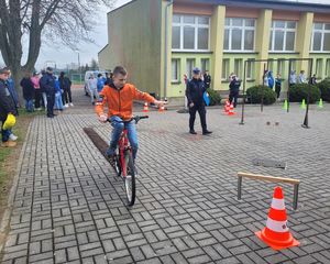 Na placu przed szkołą ustawiony tor przeszkód, uczestnik turnieju pokonuje przeszkody na rowerze.
