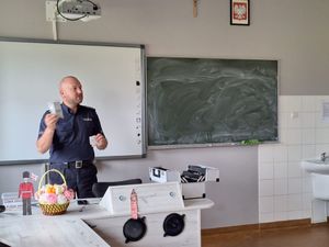 Policjant w sali lekcyjnej w szkole stoi i prezentuje atrapy narkotyków i przedmiotów służących do ich przechowywania z walizki edukacyjnej.