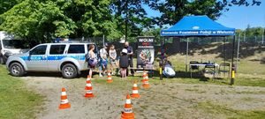 Stoisko profilaktyczne na pikniku , policjant zakłada dziecku hełm na głowę, po bokach stoi radiowóz i namiot z napisem Komenda Powiatowa Policji w Wieluniu.
