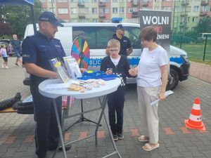 Stoisko profilaktyczne wystawione przez policjantów z wieluńskiej komendy na turnieju charytatywnym. Policjant stoi przy stoliku z upominkami obok stoi dziecko z opiekunem.