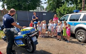 Na dziedzińcu komendy dzieci wraz z opiekunami stoją przy radiowozach i motocyklu, które prezentują policjanci.