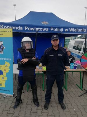 Stoisko profilaktyczne wieluńskich mundurowych. Przed namiotem policyjnym stoi policjant i uczestnik pikniku ubrany w strój wykorzystywany przez policjantów podczas zabezpieczenia imprez masowych.