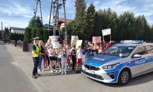 Policjanci z dziećmi z Wierzchalsa biorą udział w akcji Jabłko i Cytryna. Dzieci stoją przy radiowozie i prezentują przygotowane  na kartonach hasła profilaktyczne.
