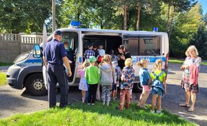 Policjant  na wewnętrznym dziedzińcu komendy pokazuje dzieciom radiowóz.