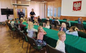 Policjanci w sali odpraw prowadzą pogadankę z dziećmi, które siedzą przy stołach.