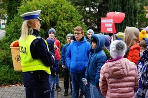 Policjantka wspólnie z dziećmi stoi na chodniku i udziela porad dotyczących bezpieczeństwa w ruchu drogowym.