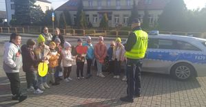 Policjant stoi z dziećmi na poboczu i udziela im wskazówek z zakresu ruchu drogowego.