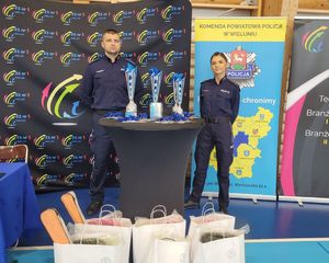 Policjanci na hali sportowej stoją przed stołem, na którym ustawione są puchary, medale i nagrody w konkursie.