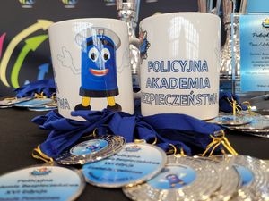 Stół z pucharami, medalami i kubkami z nadrukiem Komisarza Błyska i napisem Policyjna Akademia Bezpieczeństwa.