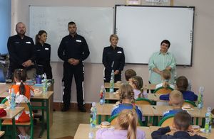Uczniowie biorący udział w konkursie siedzą w ławkach w sali. Z przodu stoją policjanci oraz wicedyrektor Zespołu Szkół numer 1 w Wieluniu.
