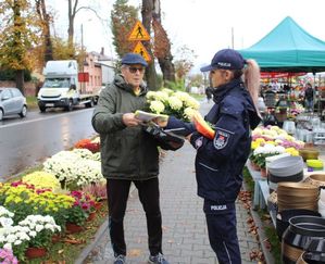 Policjantka przy cmentarzu przechodniom wręcza elementy odblaskowe i ulotki.