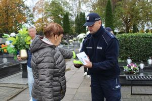 Policjant na  cmentarzu przechodniom wręcza elementy odblaskowe i ulotki.