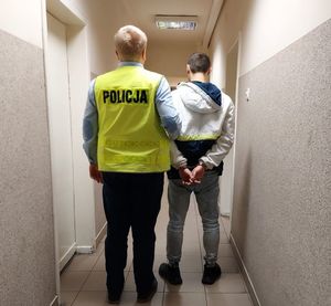 Policjant w kamizelce z napisem policja prowadzi zatrzymanego po korytarzu w budynku komendy.