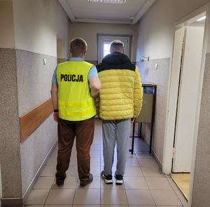 Policjant prowadzi zatrzymanego po korytarzu w budynku komendy.