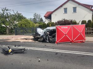 Samochód osobowy uszkodzony, który uczestniczył w wypadku drogowym stoi przy posesji. Wokół rozstawiony parawan strażacki.