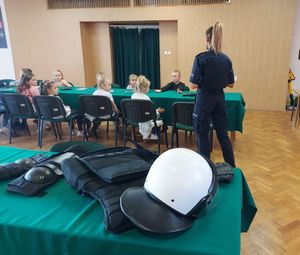 Aula komendy policji, w której przy stołach siedzą dzieci, policjantka przeprowadza prelekcję.