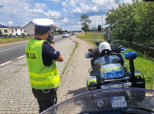 Umundurowany policjant mierzy prędkość poruszających się drogą pojazdów, obok stoi motocykl policyjny.