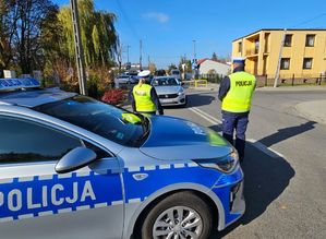 Policjanci umundurowani stoją przy drodze nadzorują ruch z boku stoi radiowóz.