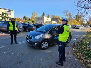 Policjanci umundurowani stoją przy drodze nadzorują ruch z boku stoi radiowóz.