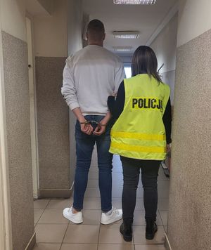 Policjantka w kamizelce z napisem Policja prowadzi zatrzymanego po korytarzu w budynku komendy.