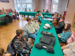 Sala w budynku komendy, przy stołach siedzą dzieci. Na stole wyłożone elementy umundurowania.