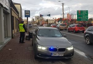 Radiowóz stoi na chodniku przy ulicy Sieradzkiej, z boku stoi policjant. Na drodze odbywał się ruch samochodów.