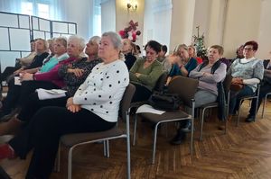 Policjant w sali Wieluńskiego Domu Kultury występuje jako prelegent. W tle prezentacja. Na krzesłach siedzą seniorzy.