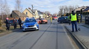 Droga, gdzie doszło do wypadku, uszkodzone samochody, które brały udział w zdarzeniu, na drodze stoi radiowóz, wóz strażacki, wokół stoją  technik kryminalistyki, strażacy i inne osoby.