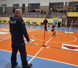 Hala sportowa uczestnik turnieju biegnie pokonując tor przeszkód. Z boku stoi policjant.