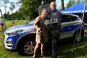 Policjanci na pikniku profilaktycznym, z uczestnikiem ubranym w hełm, trzymającym tarczę stoją na tle radiowozu.