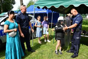 Policjanci na pikniku profilaktycznym, ubierają uczestnika w elementy umundurowania policyjnego.