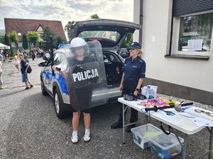 Policjantka z dziewczynką ubraną w elementy umundurowania policyjnego , w tle radiowóz.