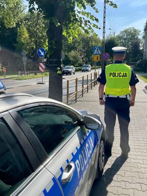 Policjant stoi przy radiowozie i  przy szkole prowadzi lustracje drogi.