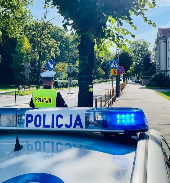 Policjant stoi przy radiowozie i  przy szkole prowadzi lustracje drogi.