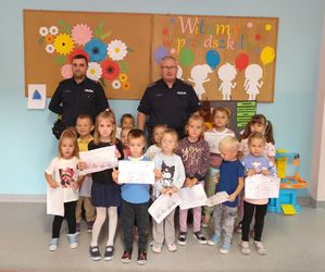 Policjanci stoją z grupą przedszkolaków w sali, dzieci pokazują rysunki.