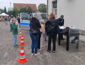 Policjantka stoi przy stole, przy którym stoją uczestnicy pikniku, w tle radiowzów.