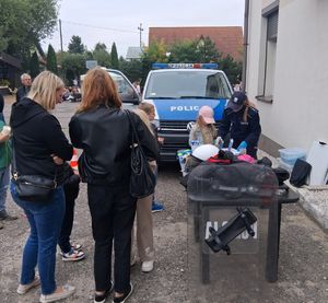 Policjantka stoi przy stole, przy którym stoją uczestnicy pikniku, w tle radiowzów