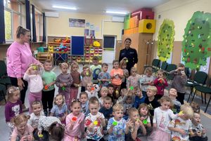 Policjanta z przedszkolakami w sali, dzieci prezentują odblaski.