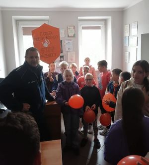 Dzieci trzymają w ręku plakat  tarcza przeciwko przemocy, jak również czerwone balony. Pośród dzieci stoi policjant.