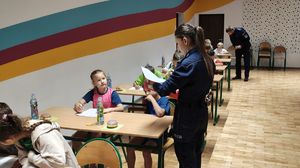 Sala lekcyjna w Zespole Szkół numer 1 w Wieluniu. Dzieci siedzą w ławkach, podchodzi do nich policjantka.