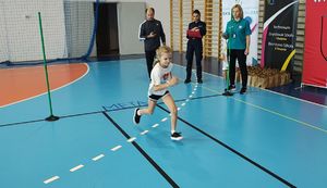 Hala sportowa. Dziewczynka biegnie na torze przeszkód. Z boku stoją sędziowie oceniający konkurencję.