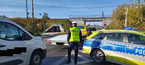 Policjant stoi przy radiowozie, z boku stoi auto uczestniczące w zdarzeniu.  przodu stoi laweta.