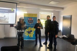 Policjanci stoją w sali w Zespole Szkół numer 1 w Wieluniu , obok stoi dziewczyna ubrana w elementy munduru wykorzystywane w trakcie zabezpieczenia przez policjantów imprez masowych.